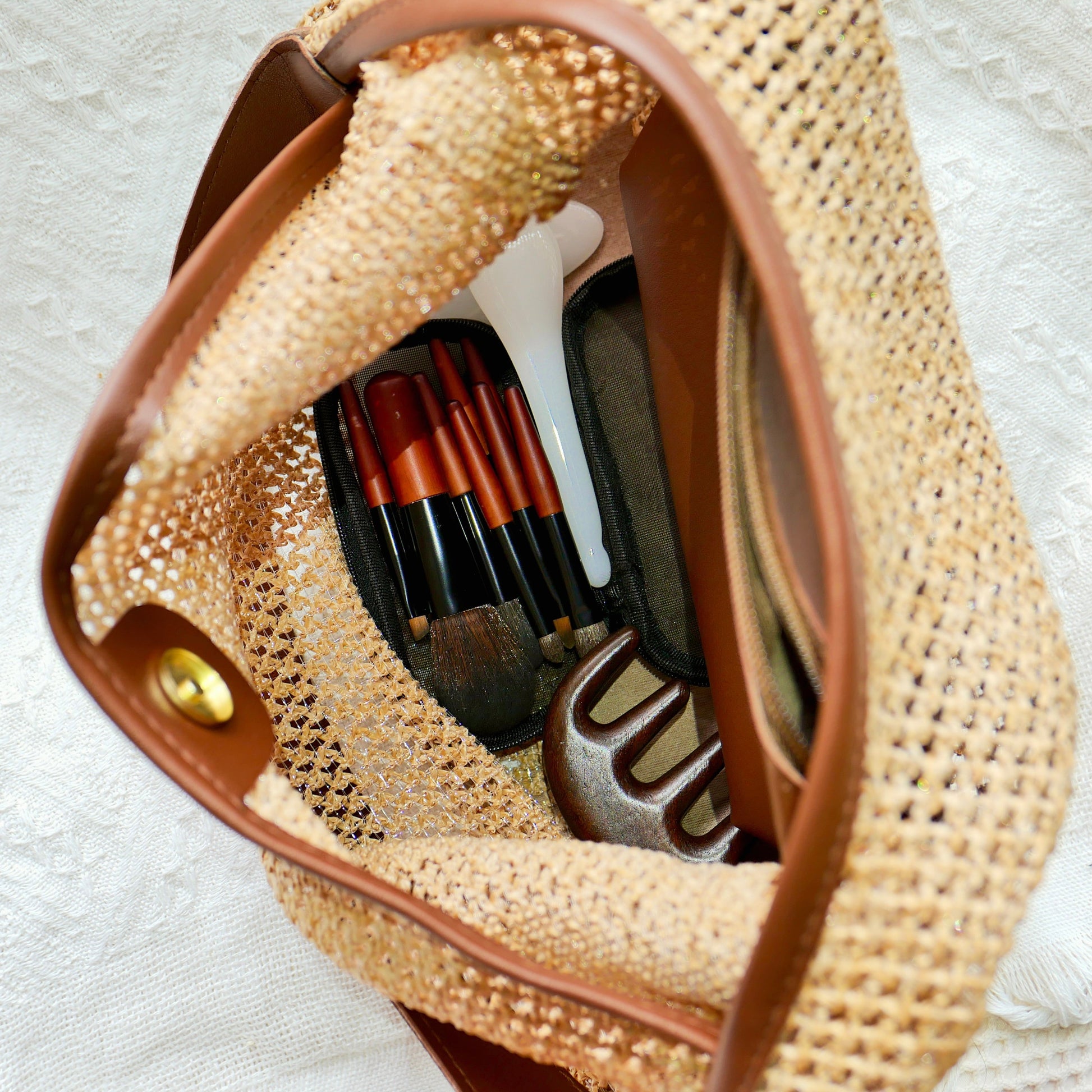 Top angle shot of a straw beach bag with mini makeup brushes, gua sha, and a wooden hair massager inside
